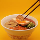 A close-up of a ramen bowl with a hand holding chopsticks about to lift a piece of fried tofu, set against a solid yellow background.