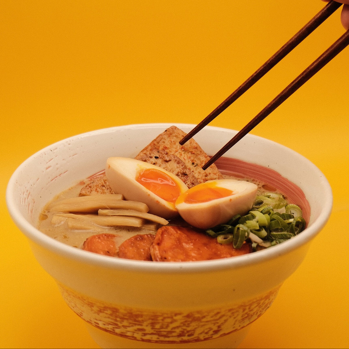 A close-up of a ramen bowl with a hand holding chopsticks about to lift a piece of fried tofu, set against a solid yellow background.
