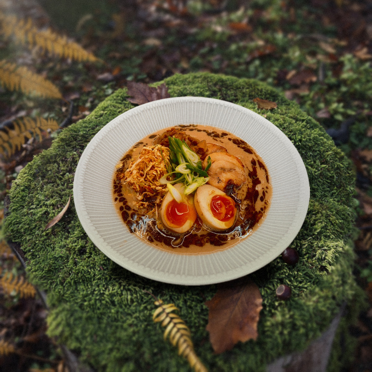 A bowl of Creamy Chicken & Mushroom Ramen on a mossy tree stump in an autumn wood, with wild mushrooms, chestnuts and fallen leaves